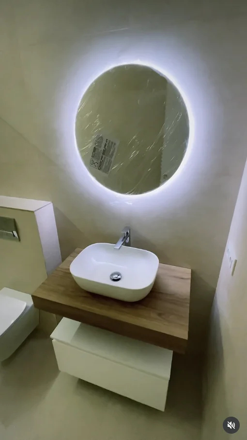 Detalle de un lavabo con espejo circular retroiluminado y encimera de madera, reforma de baño recién terminada